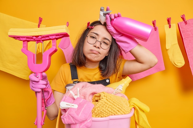 Une jeune femme au foyer asiatique surmenée et fatiguée soupire de fatigue tient une vadrouille et un détergent de nettoyage occupé à faire du travail à la maison fait le lavage ou la lessive porte des lunettes rondes et des vêtements décontractés