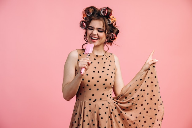 Jeune femme au foyer dans une robe à pois et bigoudis chante et danse sur fond rose.