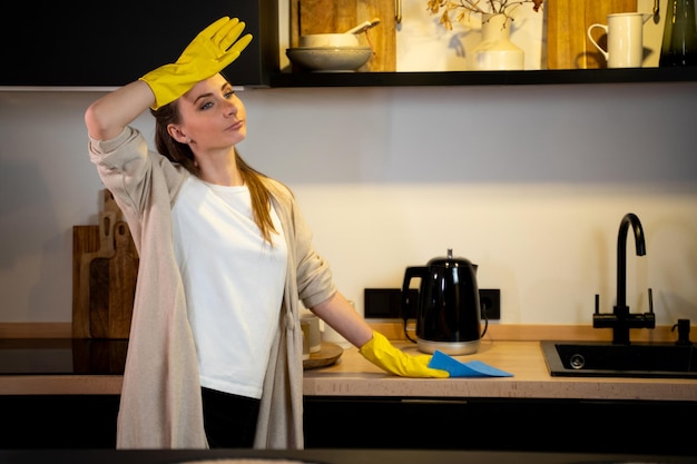 Jeune femme au foyer met des gants de protection en caoutchouc debout dans une cuisine confortable nettoie la cuisinière électrique