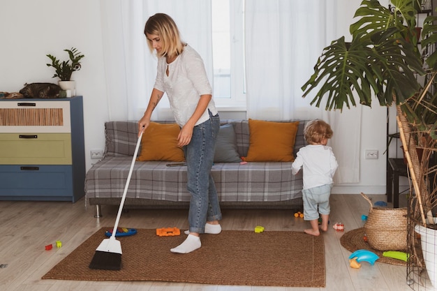 Jeune femme au foyer nettoie les jouets avec un balai avec son petit fils à la maison