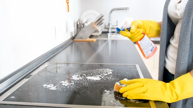 Jeune femme au foyer utilisant un spray détergent pour nettoyer la cuisinière électrique en verre dans la cuisine.