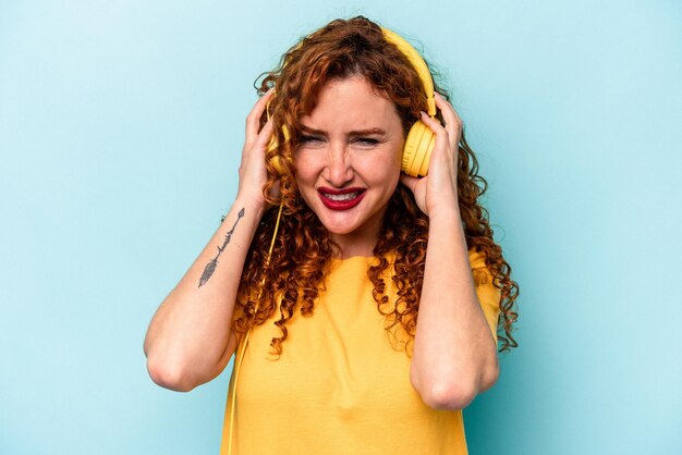 Jeune femme au gingembre écoutant de la musique isolée sur fond bleu couvrant les oreilles avec les mains