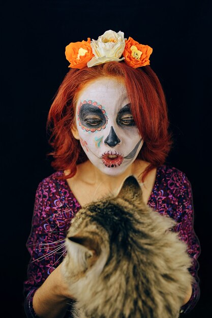 Jeune femme au jour des morts masque crâne visage art maquillage et cheveux roux jouant avec chat sur dos sombre...