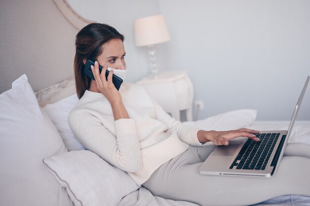 Jeune femme au lit, téléphone parlant et travaillant sur un ordinateur portable.