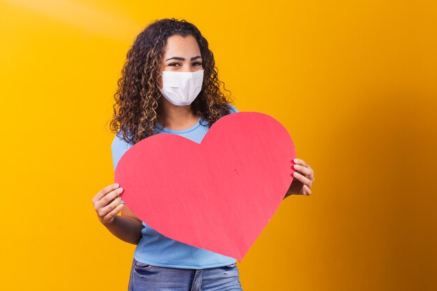 Jeune femme au masque chirurgical tenant un énorme coeur en papier avec un espace pour le texte.