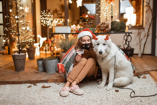 Jeune femme au masque facial célébrant avec un chien les vacances du Nouvel An à la maison. Concept de quarantaine et d'auto-isolement pendant l'épidémie en vacances