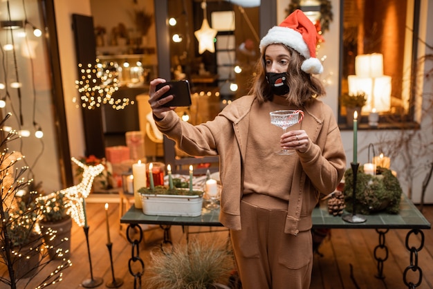 Jeune femme au masque facial célébrant seule les vacances du Nouvel An à la maison, ayant un appel vidéo au téléphone avec des amis. Concept de quarantaine et d'auto-isolement pendant l'épidémie en vacances