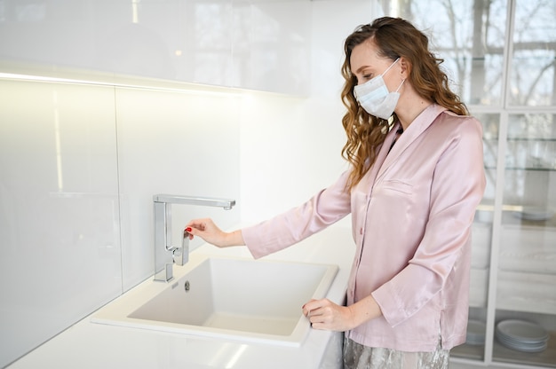 Jeune femme au masque facial se laver les mains pour la prévention du virus corona, l'hygiène pour arrêter la propagation du coronavirus. Évitez de contaminer le virus Corona Covid-19 en quarantaine à domicile dans un intérieur de cuisine blanc