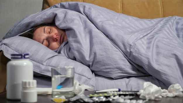 Une jeune femme au nez qui coule tousse fortement allongée sur un canapé sous une couverture confortable près d'une table avec des médicaments prescrits dans le salon.