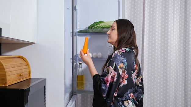 Jeune femme au régime affamée en robe sombre avec imprimé floral ouvre la porte d'un réfrigérateur moderne pour prendre un gâteau sucré dans la cuisine la nuit vue rapprochée