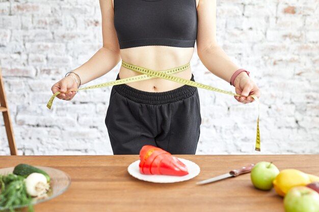 Jeune femme au régime, elle mesure sa taille avec un ruban à mesurer doux, des légumes frais sur la table