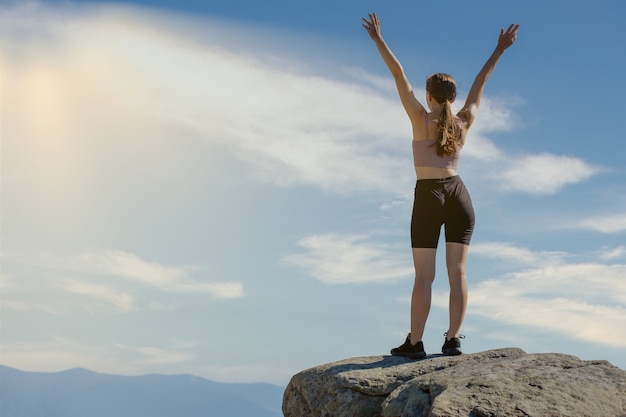 La jeune femme au sommet de la montagne leva les mains sur fond de ciel bleu.