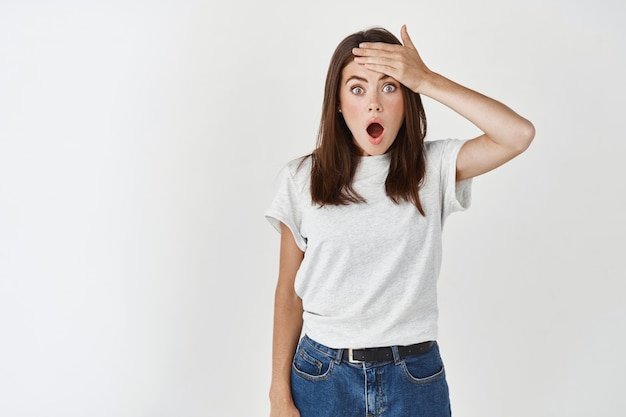 Jeune femme au visage surpris regardant devant, la mâchoire tombante et gifle le front avec admiration, debout émerveillée contre le mur blanc