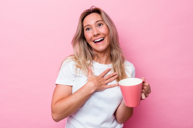 Jeune femme australienne tenant une tasse rose isolée sur fond rose éclate de rire en gardant la main sur la poitrine.