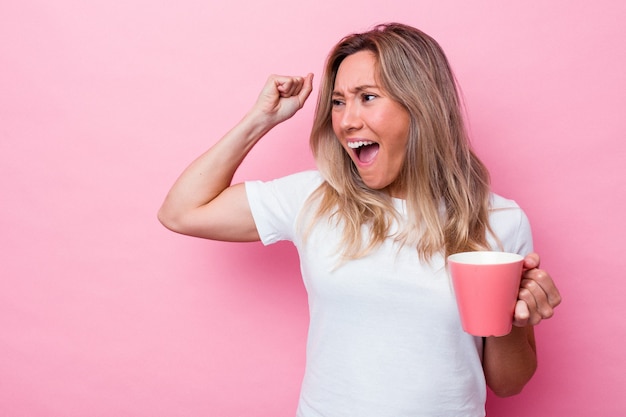 Jeune femme australienne tenant une tasse rose isolée sur fond rose levant le poing après une victoire, concept gagnant.