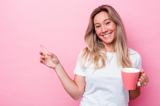 Jeune femme australienne tenant une tasse rose isolée sur fond rose souriante et pointant de côté, montrant quelque chose dans un espace vide.