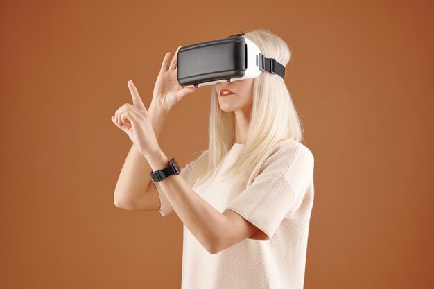 Jeune femme aux cheveux blonds curieuse moderne en tshirt debout sur fond marron et touchant un tableau invisible tout en utilisant un appareil de réalité virtuelle