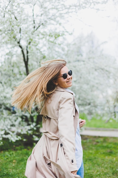 Une jeune femme aux cheveux blonds profite d'un jardin printanier fleuri Travel spring break Style à la mode Une femme portant des lunettes de soleil et un trench beige traverse un parc fleuri Mise au point de sélection