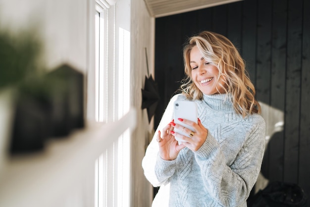 Jeune femme aux cheveux bouclés blonds en pull gris utilisant un téléphone portable dans les mains près de la fenêtre de la maison