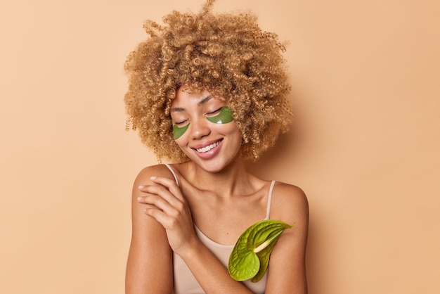 Une jeune femme aux cheveux bouclés et doux garde les yeux fermés touche l'épaule applique des patchs de collagène vert pour éliminer les cernes isolés sur fond beige profite de la routine de soins de la peau