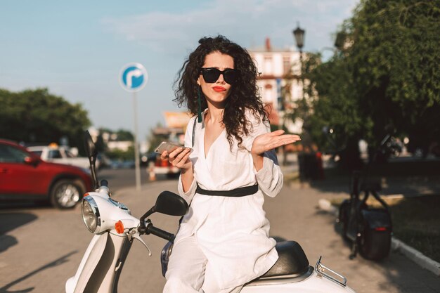 Jeune femme aux cheveux bouclés noirs en costume blanc et lunettes de soleil assis sur un cyclomoteur blanc avec un téléphone portable à la main dans la rue avec vue sur la ville en arrière-plan