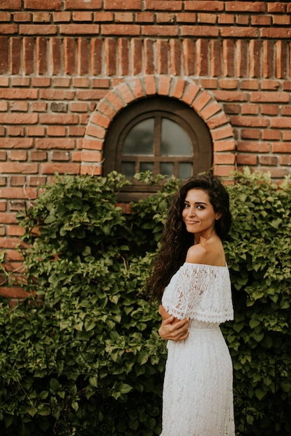 Une jeune femme aux cheveux bouclés se tient dans un beau jardin vêtue d'une robe blanche fluide