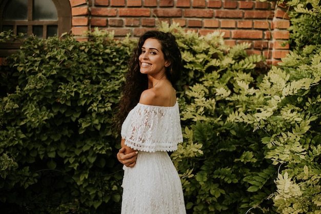 Une jeune femme aux cheveux bouclés se tient dans un beau jardin vêtue d'une robe blanche fluide