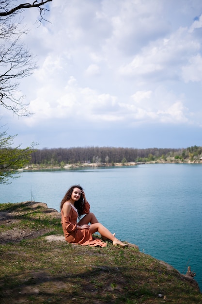 Une jeune femme aux cheveux bouclés et un sourire sur son visage est assise dans un pré vert sur fond de lac bleu. Chaude journée d'été, fille heureuse, émotions de joie