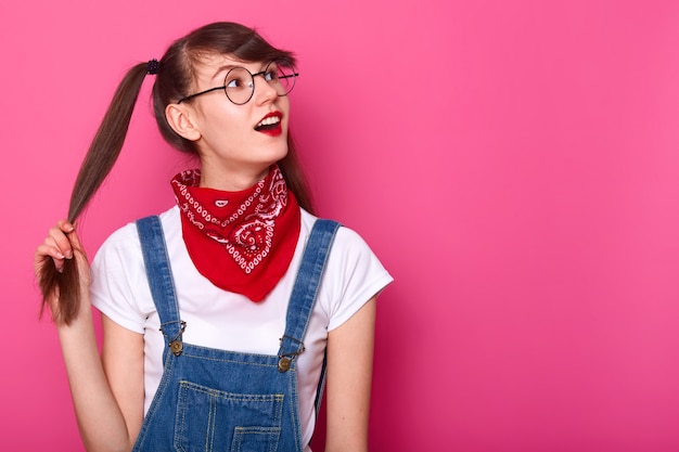 Jeune femme aux cheveux bruns brillants en bonne santé, tient sa queue de cheval à la main. Écolière intéressée par quelque chose