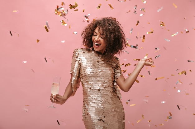 Jeune femme aux cheveux bruns courts en robe élégante brillante posant avec un verre de vin et des confettis sur un mur isolé.