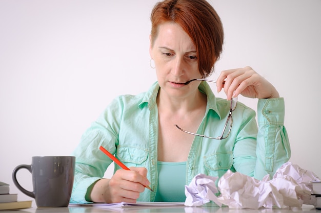 Jeune femme aux cheveux courts au bureau écrit pensivement au crayon rouge Papier froissé sur le bureau