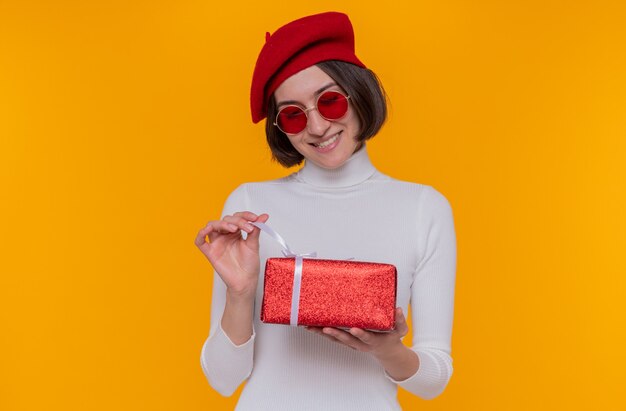 Jeune femme aux cheveux courts en col roulé blanc portant un béret et des lunettes de soleil rouges tenant un cadeau heureux et joyeux va ouvrir présent souriant joyeusement