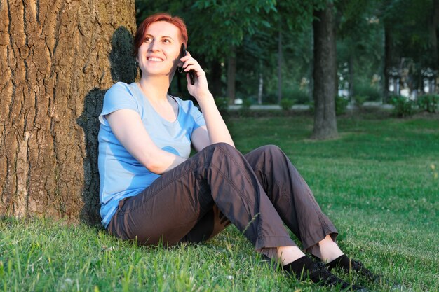 Jeune femme aux cheveux courts est assise sur l'herbe près d'un grand arbre et parle sur son téléphone portable