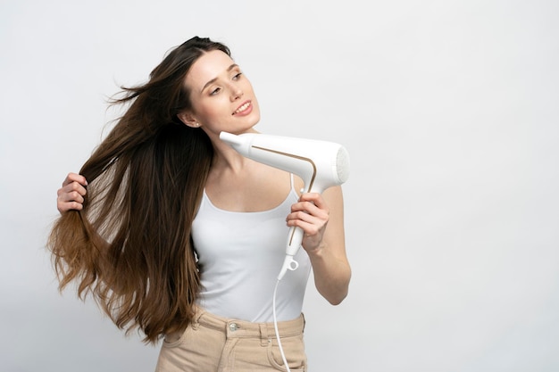 une jeune femme aux cheveux lisses et soyeux brillants qui regarde rêveusement de côté en utilisant un sèche-cheveux