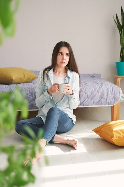 Une jeune femme aux cheveux longs est assise par terre à la maison et boit du café