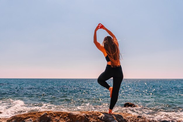 Jeune femme aux cheveux longs instructeur de fitness qui s'étend avant le pilates sur un tapis de yoga près de la mer par une journée ensoleillée concept de routine de yoga fitness féminin