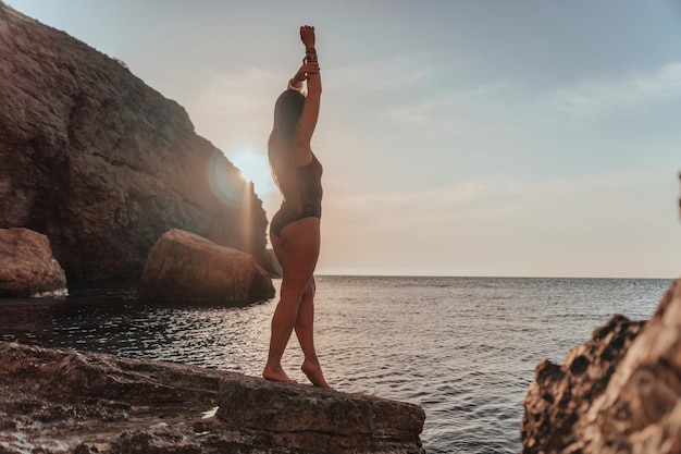 Jeune femme aux cheveux longs en maillot de bain noir et braclets de style bohème pratiquant à l'extérieur sur un tapis de yoga au bord de la mer au coucher du soleil Routine de fitness yoga pour femmes Harmonie et méditation d'un mode de vie sain