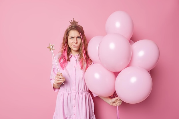 Une jeune femme aux cheveux longs malheureuse et frustrée tient un tas de ballons gonflés et une baguette magique en colère contre quelqu'un qui porte une robe et des poses de couronne sur fond rose n'aime pas quelque chose