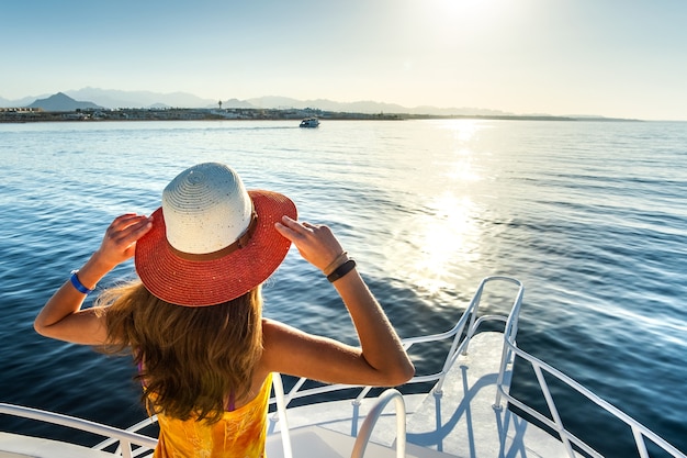 Jeune femme aux cheveux longs portant une robe jaune et un chapeau de paille debout sur un pont de yacht blanc bénéficiant d'une vue sur l'eau de mer bleue.