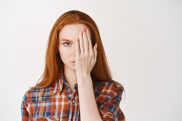 Jeune femme aux cheveux longs rouges et à la peau pâle, couvrant la moitié du visage et regardant sérieusement à l'avant, debout sur un mur blanc