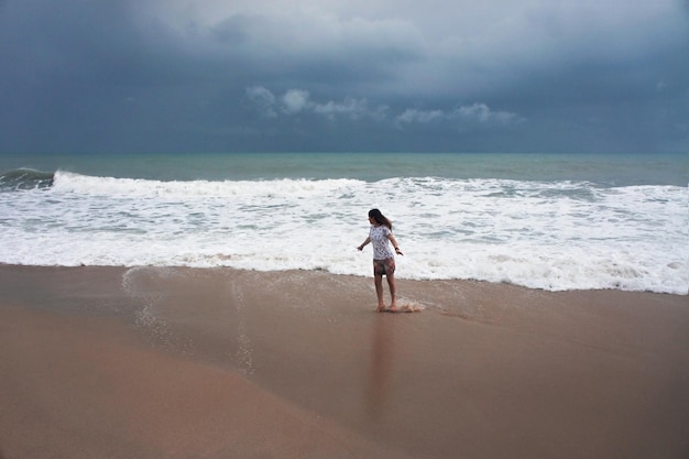 Jeune femme aux cheveux longs sautant dans la mousse des vagues de la mer pendant la tempête sur fond de ciel pluvieux