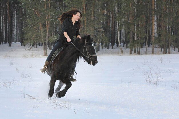 Jeune femme aux cheveux noirs sur un cheval bai en forêt d'hiver, téléobjectif