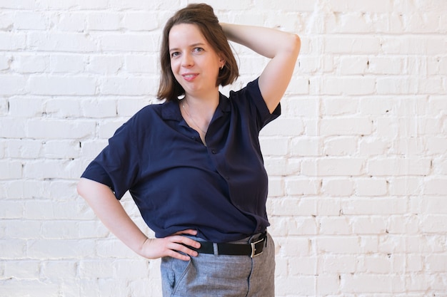 Jeune femme aux cheveux noirs jusqu'aux épaules en pantalon et chemise bleu marine se dresse contre un mur de briques blanches. La femme pose, sourit et met sa main derrière sa tête.