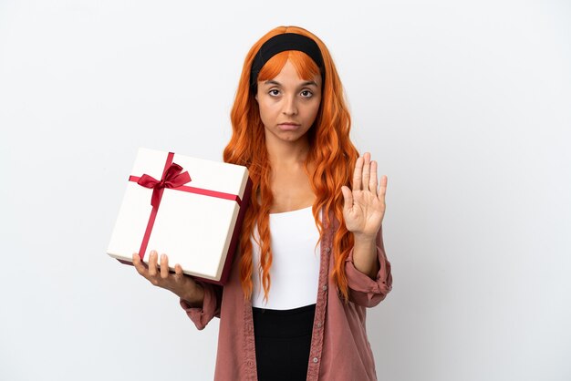 Jeune femme aux cheveux orange tenant un cadeau isolé sur fond blanc faisant un geste d'arrêt