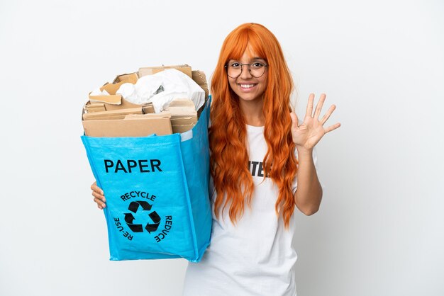 Jeune femme aux cheveux orange tenant un sac de recyclage plein de papier à recycler isolé sur fond blanc en comptant cinq avec les doigts