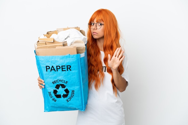 Jeune femme aux cheveux orange tenant un sac de recyclage plein de papier à recycler isolé sur fond blanc faisant un geste d'arrêt et déçu