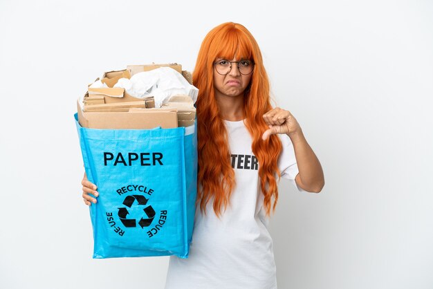 Jeune femme aux cheveux orange tenant un sac de recyclage plein de papier à recycler isolé sur fond blanc montrant le pouce vers le bas avec une expression négative