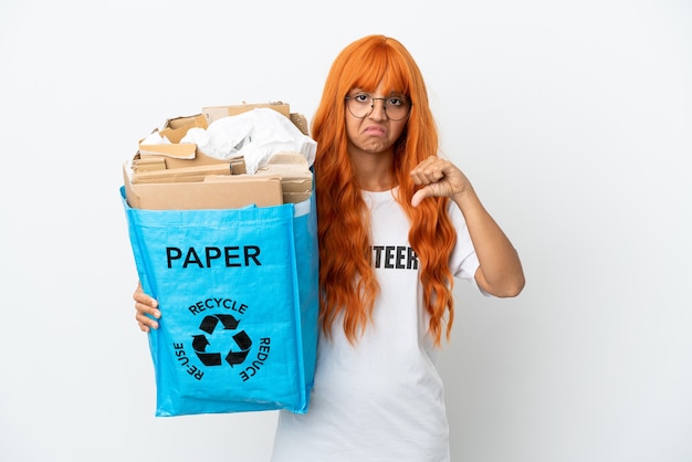 Jeune femme aux cheveux orange tenant un sac de recyclage plein de papier à recycler isolé sur fond blanc montrant le pouce vers le bas avec une expression négative