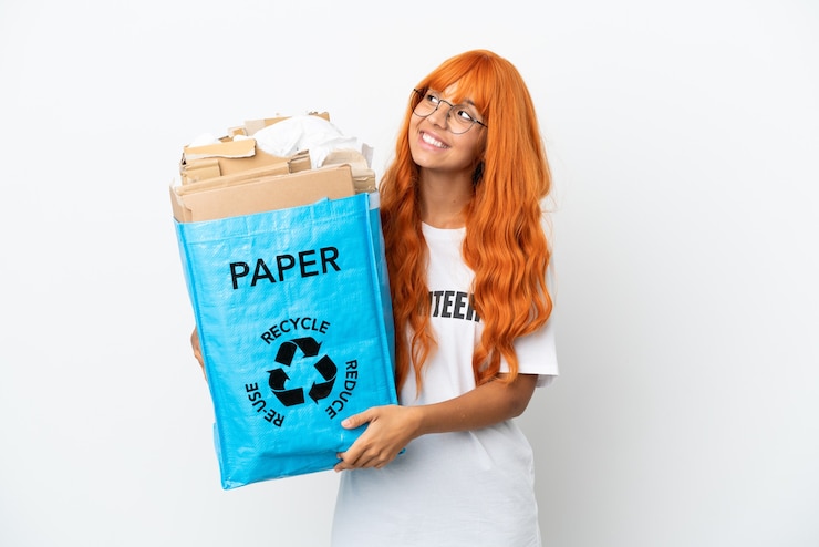 Photo jeune femme aux cheveux orange tenant un sac de recyclage plein de papier à recycler isolé sur fond blanc en pensant à une idée tout en levant les yeux
