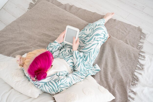 Jeune femme aux cheveux roses et blancs colorés lisant un appareil numérique de livre électronique allongé dans son lit à la maison